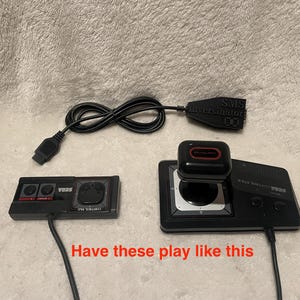 May include: A black Sega Genesis console with a black joystick and a black controller. The console has a red and black Sega logo and the text "SEGA GENESIS" printed on it. The joystick has a red button and a black base. The controller has a red button and a black base. The console is connected to the joystick and controller with black cables.