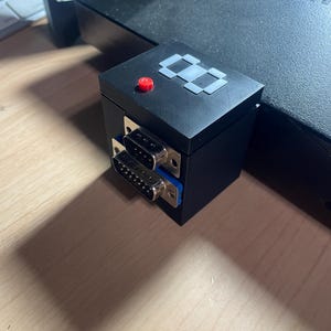 May include: Black electronic device with two ports and a red button on top. The device has a geometric design on the top and is connected to a larger black component. The image is taken from a low angle.