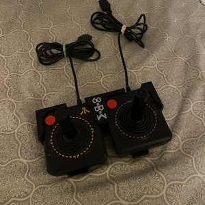 May include: Two black Atari joysticks with red buttons and a gold trim. The joysticks are connected to a black base with the text "8-B-3" and "TOP". The joysticks have black cords with rubber bands.