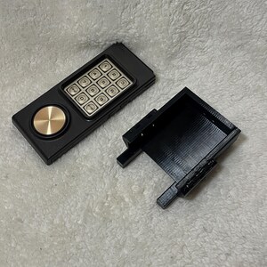 May include: A black keypad device with a gold-colored circular button and a separate black rectangular case. The keypad has silver buttons with numbers and function labels. The device is on a white, textured surface.