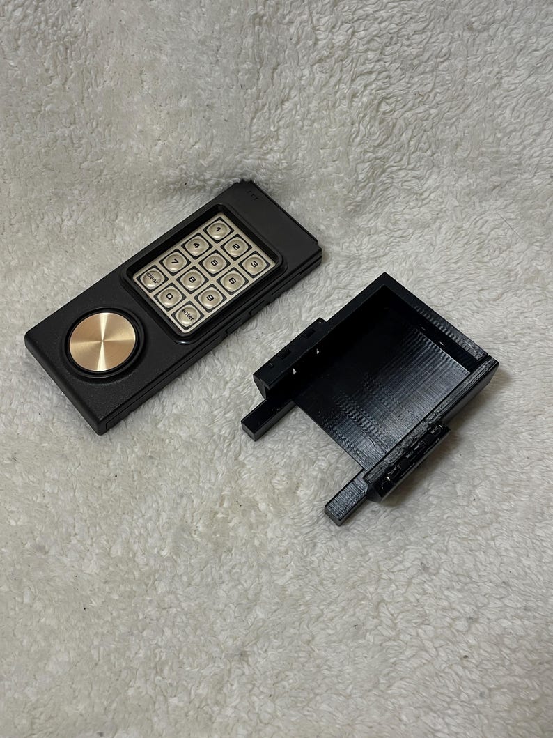 May include: A black keypad device with a gold-colored circular button and a separate black rectangular case. The keypad has silver buttons with numbers and function labels. The device is on a white, textured surface.