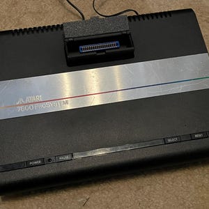 May include: A black Atari 7800 ProSystem video game console with a silver and black stripe across the front. The console has a slot for a game cartridge, and buttons for power, pause, select, and reset.