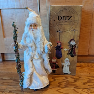 May include: A white, plush Santa Claus marionette with a long white beard and glasses. He is holding a green and gold twig with bells. The marionette is hanging from strings and is in front of a box that says "Marionettes DITZ designs by The Hen House, Inc."