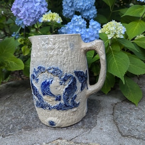 May include: A beige stoneware pitcher with a textured surface and a blue floral design. The pitcher has a large handle and a spout. The blue design is centered on the front of the pitcher. The pitcher is sitting on a stone surface with blue and purple flowers in the background.