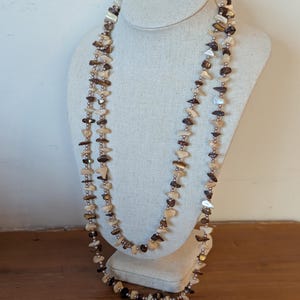 May include: A long, layered necklace displayed on a mannequin. The necklace features a mix of light brown, dark brown, and cream-colored shell fragments strung together with small silver beads. The necklace is a natural, earthy accessory.