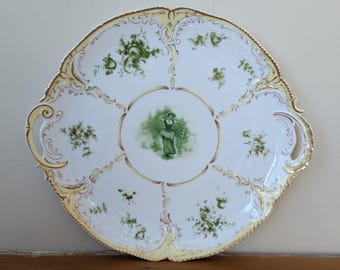 May include: A white porcelain serving platter with a scalloped edge and gold trim. The platter features green floral designs and a central image of a person. The handles are gold and the edge is a pale yellow.