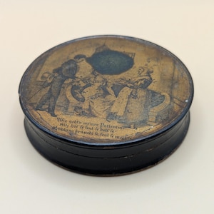 May include: A small, round, antique box with a black base and a detailed lid. The lid features a sepia-toned illustration of figures and text in German. The box appears to be made of metal or a similar material.