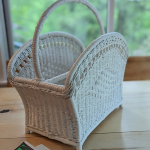 May include: White wicker magazine holder with a handle. The holder has a woven design and is in the shape of a basket.