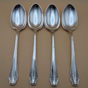 May include: Four silver spoons arranged in a row. Each spoon has an oval bowl and a long, slender handle with a decorative floral pattern at the base. The spoons are reflective, with a polished finish.