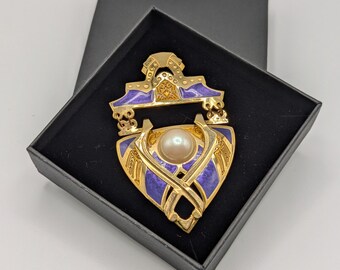 May include: A gold-toned pendant with a purple enamel design and a white pearl in the center. The pendant is in a black velvet box.