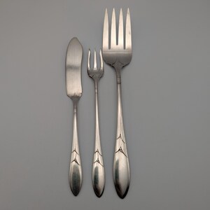 May include: A set of three silver-colored utensils: a butter knife, a small fork, and a large fork. The utensils have a teardrop-shaped handle with a decorative design near the top. The background is a solid gray.