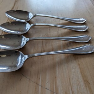 May include: Four silver spoons with ornate handles. The spoons are arranged in a row, with the handles pointing to the right. The spoons are polished and shiny.