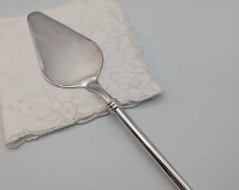 May include: Silver cake server with a long, thin handle and a rounded, slightly pointed blade. The handle has a decorative, ridged design.