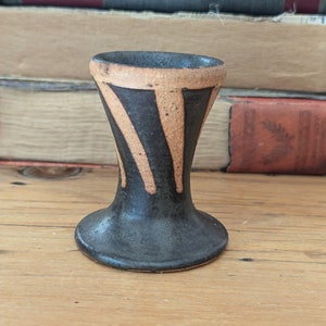 May include: A small, black and tan ceramic vase with a flared base and a narrow opening. The vase has a striped pattern.
