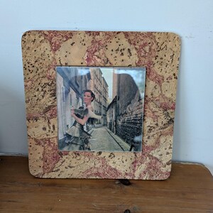 May include: A square cork photo frame with a picture of a woman in a dress standing on a cobblestone street. The frame has a natural cork color with red and brown marbling.