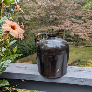 May include: A dark brown stoneware jug with a handle, sitting on a wooden railing. The jug has a slightly glazed finish and a rustic appearance.