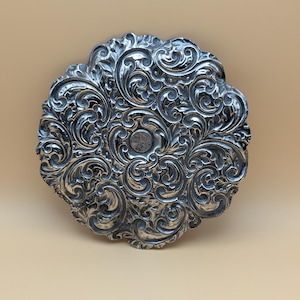 May include: A silver-toned, circular decorative plate with an ornate, swirling floral pattern. The plate has a scalloped edge and a small, circular indentation in the center. The design is highly detailed and raised, creating a textured surface.