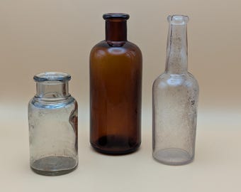 May include: Three antique glass bottles of varying sizes and shapes. The bottles are clear, amber, and brown. The bottles have a vintage aesthetic, with signs of wear and age. The bottles are displayed against a neutral background.