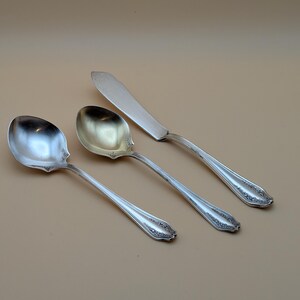 May include: Three pieces of silver silverware are displayed against a neutral background. The set includes two spoons and a butter knife. The silverware has decorative detailing on the handles.