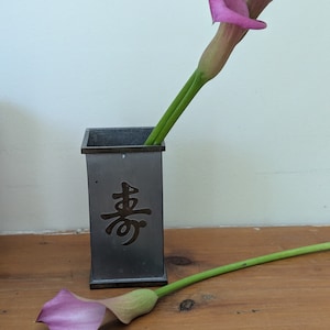 May include: Two pink calla lilies in a gray metal vase with the Chinese character for "joy" inscribed on the side. The vase is sitting on a wooden surface.