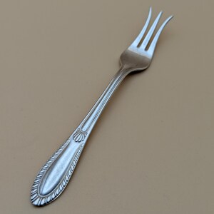 May include: A silver fork with three tines, featuring a decorative handle with a detailed pattern. The fork is angled against a neutral background, showcasing its elegant design. This utensil is suitable for formal dining.