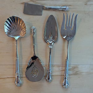 May include: A collection of antique silver serving utensils, including a cleaver, a shell-shaped ladle, a serving spoon, a pie server, and a large fork. The utensils are arranged on a light wood surface, showcasing their ornate handles and polished finish.