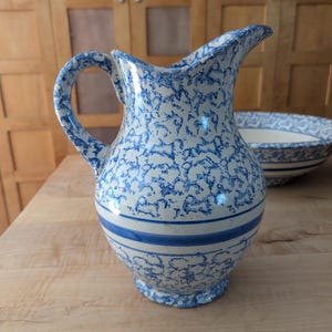May include: A white ceramic pitcher with a blue and white floral pattern. The pitcher has a wide mouth and a handle. It has a blue stripe around the middle.