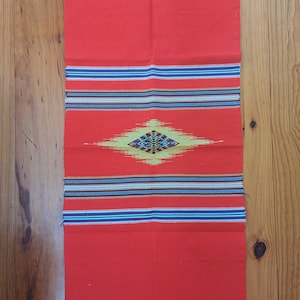 May include: Red woven fabric with a diamond pattern in the center. The diamond is made of yellow, blue, and black threads. The fabric has a fringe border.