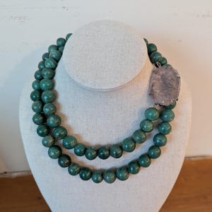 May include: A two-strand necklace with large, round, teal-green beads. A rectangular, carved, lavender-gray pendant hangs from the necklace. The necklace is displayed on a white mannequin bust against a neutral background.
