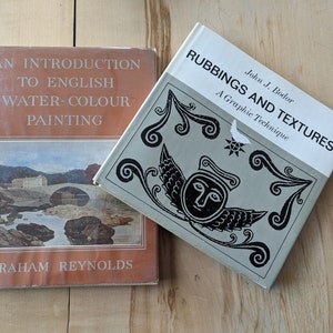 May include: Two books on a wooden surface. The book on the left is titled "An Introduction to English Water-Colour Painting" by Graham Reynolds. The book on the right is titled "Rubbings and Textures: A Graphic Technique" by John J. Bodor.
