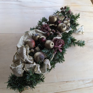May include: A gold and burgundy Christmas garland with faux fruit, berries, and roses. The garland is made of green pine branches and has a gold ribbon.