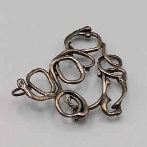 May include: A silver abstract brooch with a unique, organic design. The brooch is made of twisted and curved metal, creating a series of interconnected loops and circles.