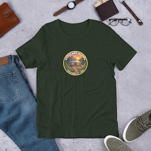 May include: Dark green t-shirt with a circular graphic featuring a cabin in a landscape with the text "BASECAMP BLUFFS GUILFORD, CT - USA". The shirt is made of a soft material and is suitable for casual wear.