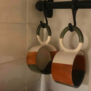 May include: Two ceramic mugs with a green, white, and orange color scheme hang from hooks on a black metal rack. The mugs have a unique shape with a double loop handle.
