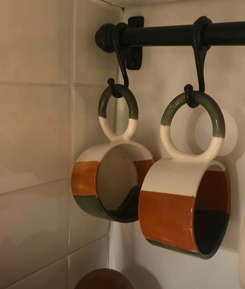 May include: Two ceramic mugs with a green, white, and orange color scheme hang from hooks on a black metal rack. The mugs have a unique shape with a double loop handle.