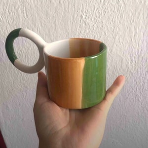 May include: A white ceramic mug with a unique loop handle. The mug is painted with three vertical stripes in green, orange, and brown.