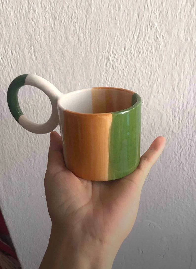 May include: A white ceramic mug with a unique loop handle. The mug is painted with three vertical stripes in green, orange, and brown.