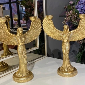 May include: Two gold candle holders shaped like a winged woman with outstretched arms. Each holder has two candle cups.