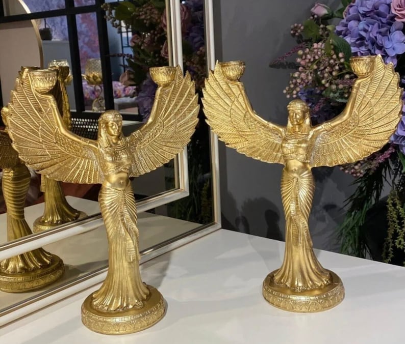 May include: Two gold candle holders shaped like a winged woman with outstretched arms. Each holder has two candle cups.