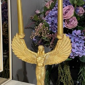 May include: Gold candle holder featuring a winged goddess figure with a flowing gown. The holder has two candle slots and a decorative base.