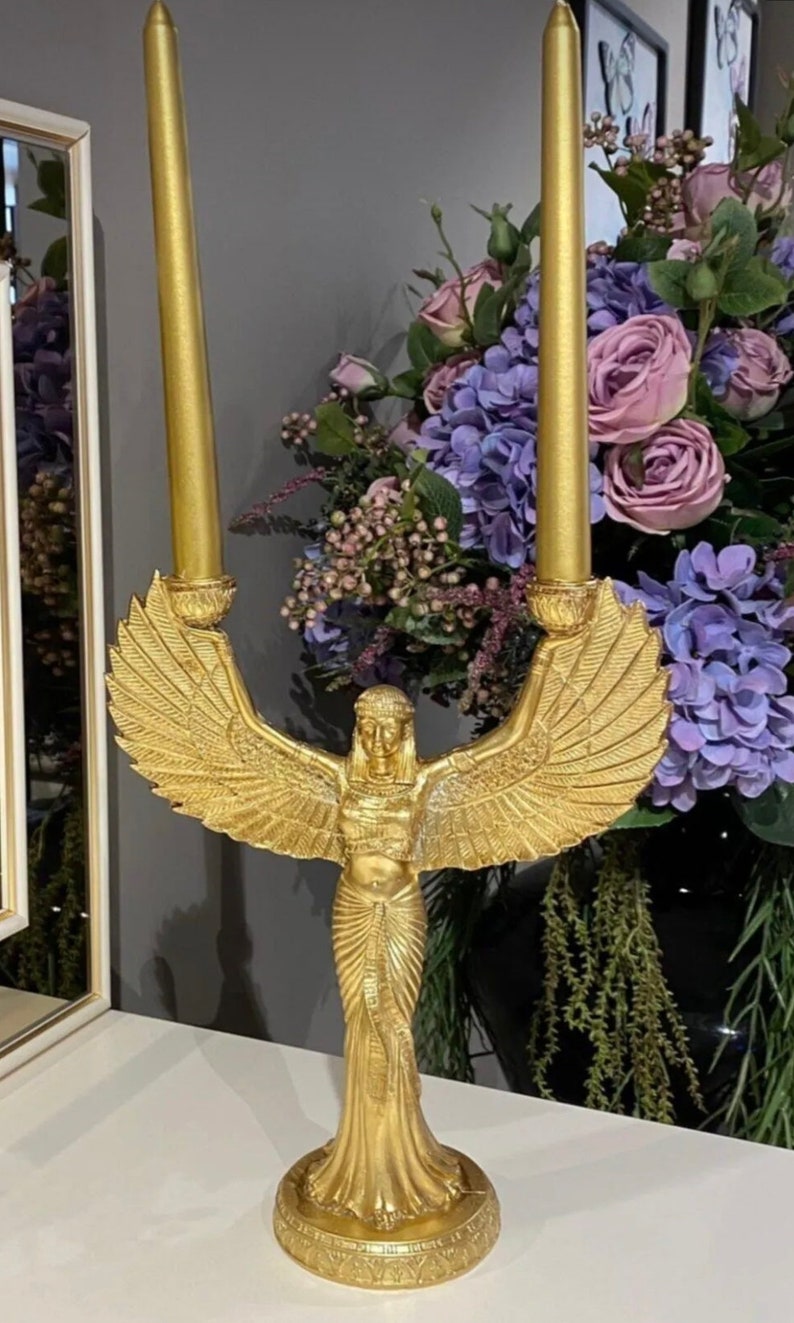 May include: Gold candle holder featuring a winged goddess figure with a flowing gown. The holder has two candle slots and a decorative base.