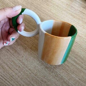May include: A white ceramic mug with a unique double handle. The mug is painted with three horizontal stripes in green, beige, and white.