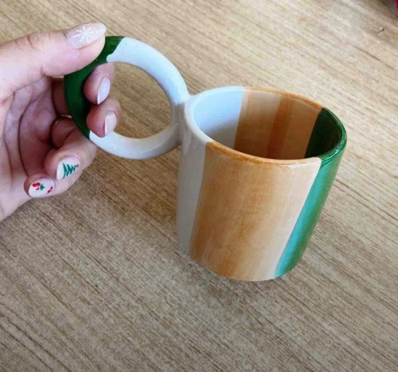May include: A white ceramic mug with a unique double handle. The mug is painted with three horizontal stripes in green, beige, and white.