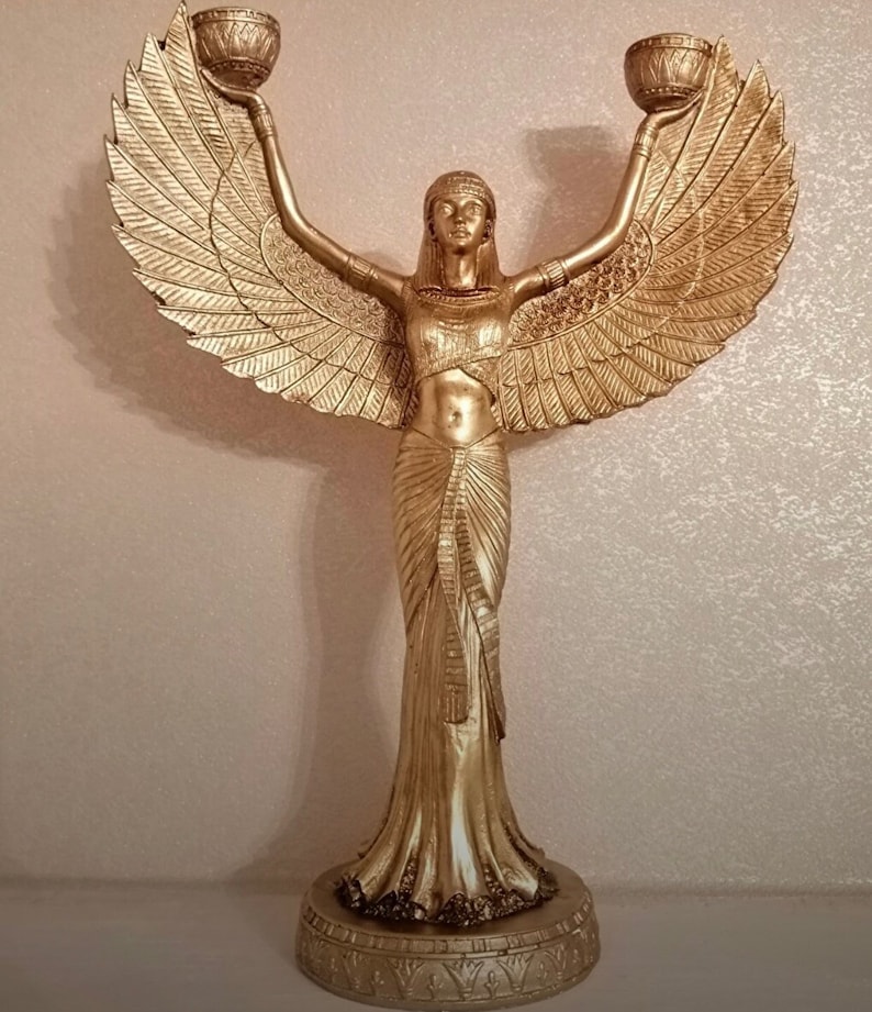 May include: A gold-colored statue of a winged woman holding two candle holders. The statue is standing on a base with an intricate design.