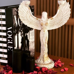 May include: White ceramic candle holder with a winged figure design. The figure is wearing a long, flowing dress and has a crown on her head. The candle holder is sitting on a table with a magazine and candles.