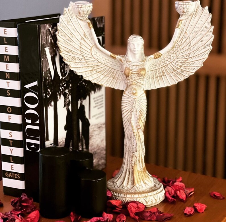 May include: White ceramic candle holder with a winged figure design. The figure is wearing a long, flowing dress and has a crown on her head. The candle holder is sitting on a table with a magazine and candles.