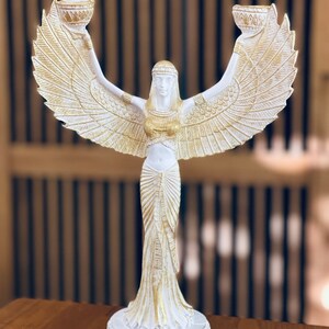 May include: White and gold decorative candle holder featuring a winged female figure with outstretched arms holding two candle cups. The figure is wearing a flowing gown and has a crown on her head.
