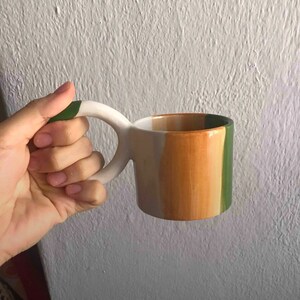 May include: A white ceramic mug with a green, orange, and white striped design. The mug has a unique, curved handle.
