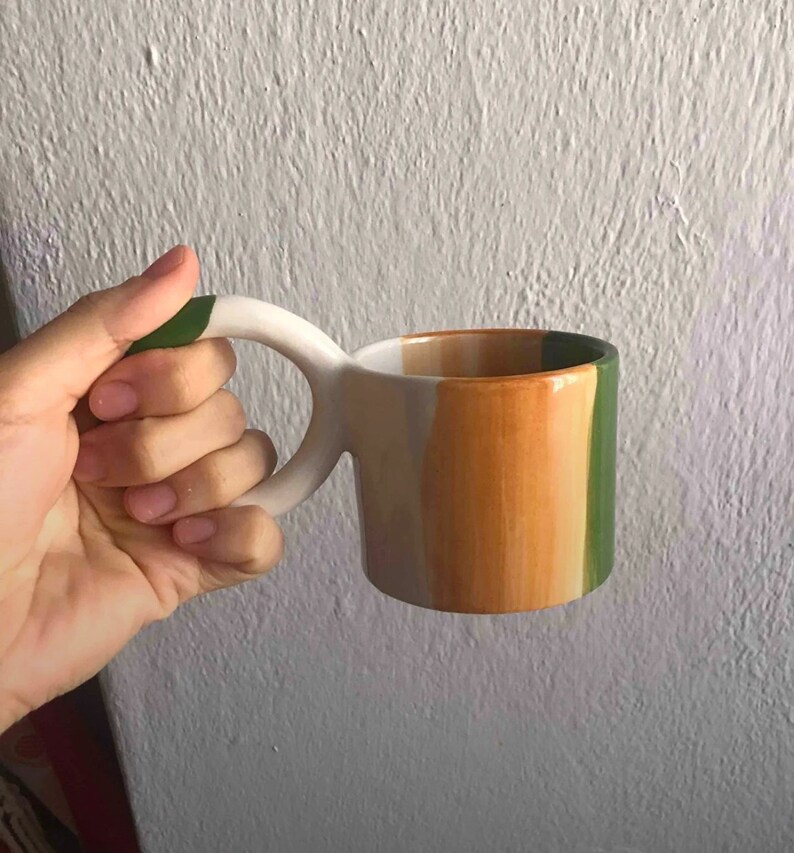 May include: A white ceramic mug with a green, orange, and white striped design. The mug has a unique, curved handle.