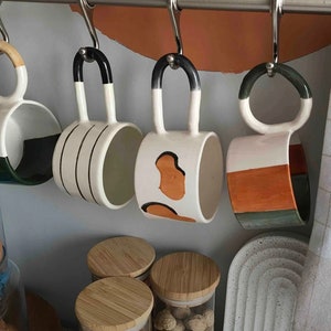 May include: Four ceramic mugs with different patterns hang on a metal rack. The mugs are white with black, orange, and green accents. The mugs are hanging on a metal rack with hooks.